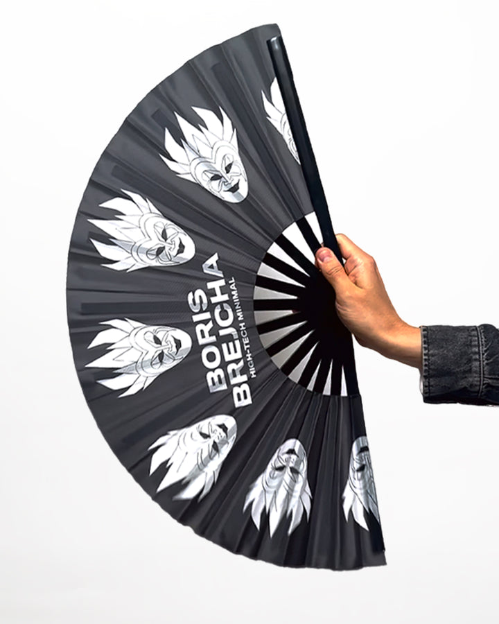 Hand holds open a black folding hand fan with silver stylized face designs and "Boris Brejcha High-Tech Minimal" printed on it. 
